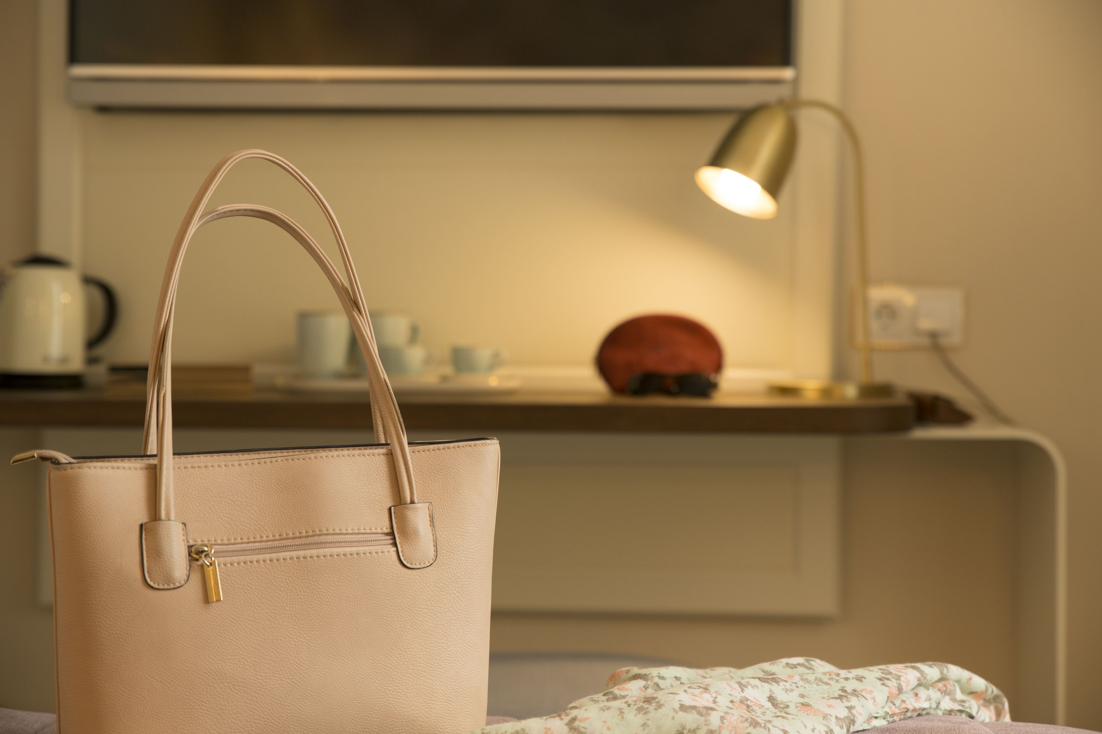 A beige handbag on a bed with a floral blanket, a lamp, and a red object on a bedside table.