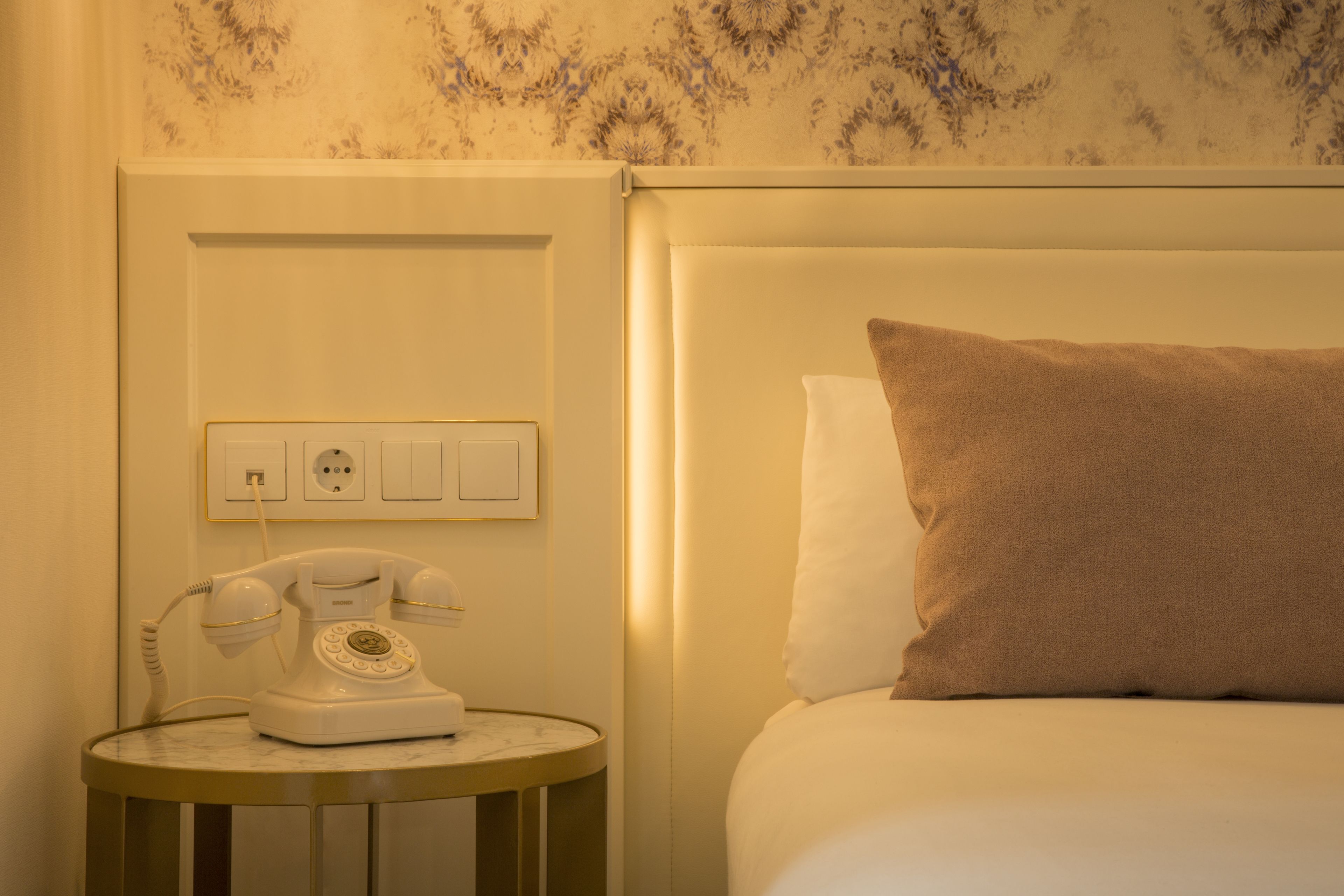 A hotel room with a bed, a bedside table with a rotary phone, and a wall socket.