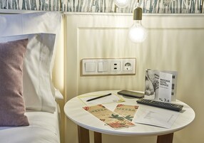 A neatly arranged hotel room with a bed, a bedside table with a lamp, a remote control, a smartphone, and a brochure.