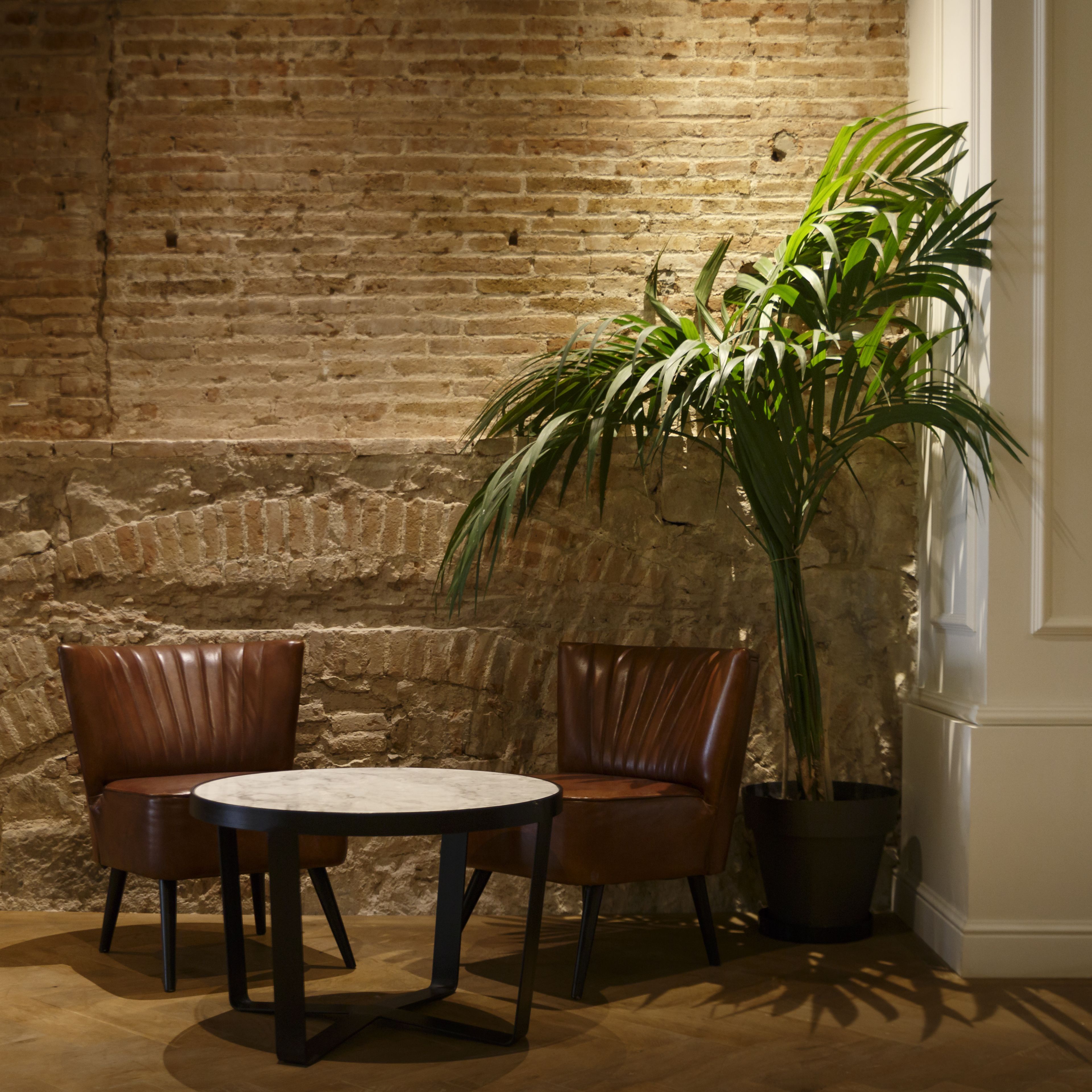 Una habitación con pared de ladrillo, una planta en maceta, dos sillones de cuero y una mesa redonda.
