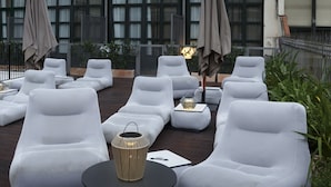 Un área de lounge en la azotea con sillones y mesas blancas, un farol decorativo y vistas de un edificio con ventanas grandes y un balcón.