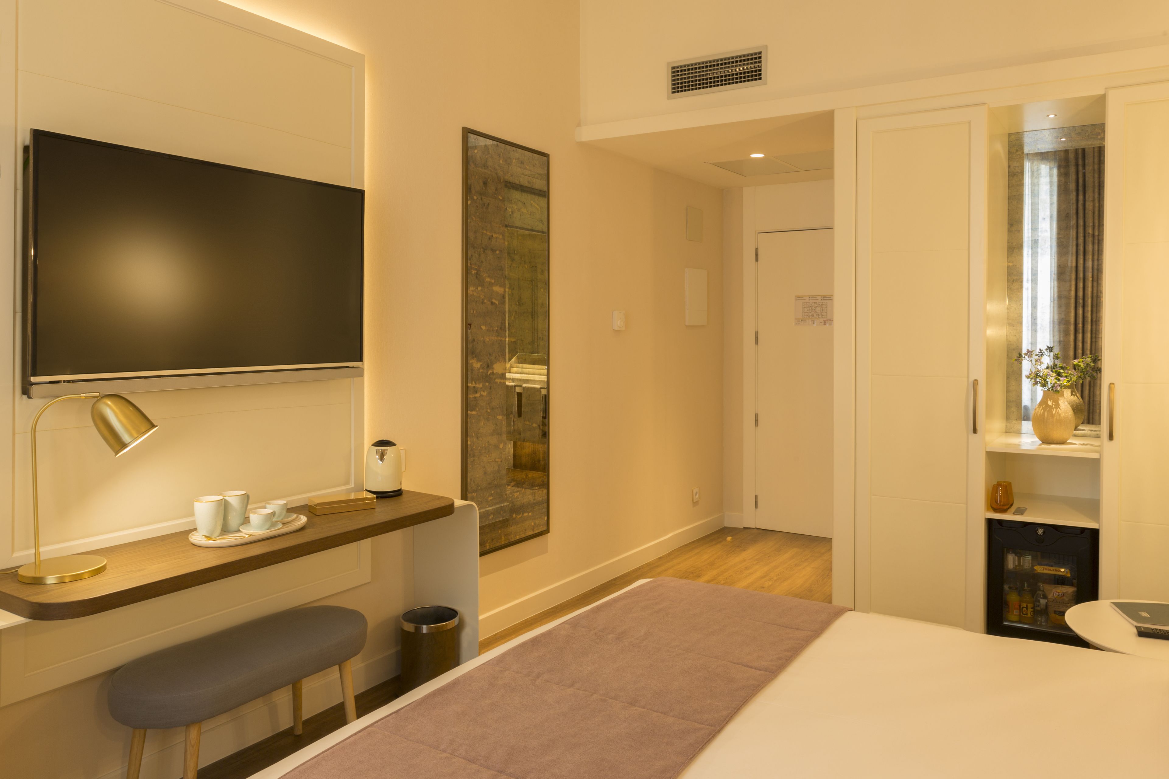 A modern hotel room with a flat-screen TV, a desk with a lamp, a bench, and a bed.