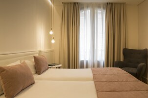Quarto de hotel com uma cama grande, duas almofadas, mesa de cabeceira, uma cadeira e janela com cortinas.