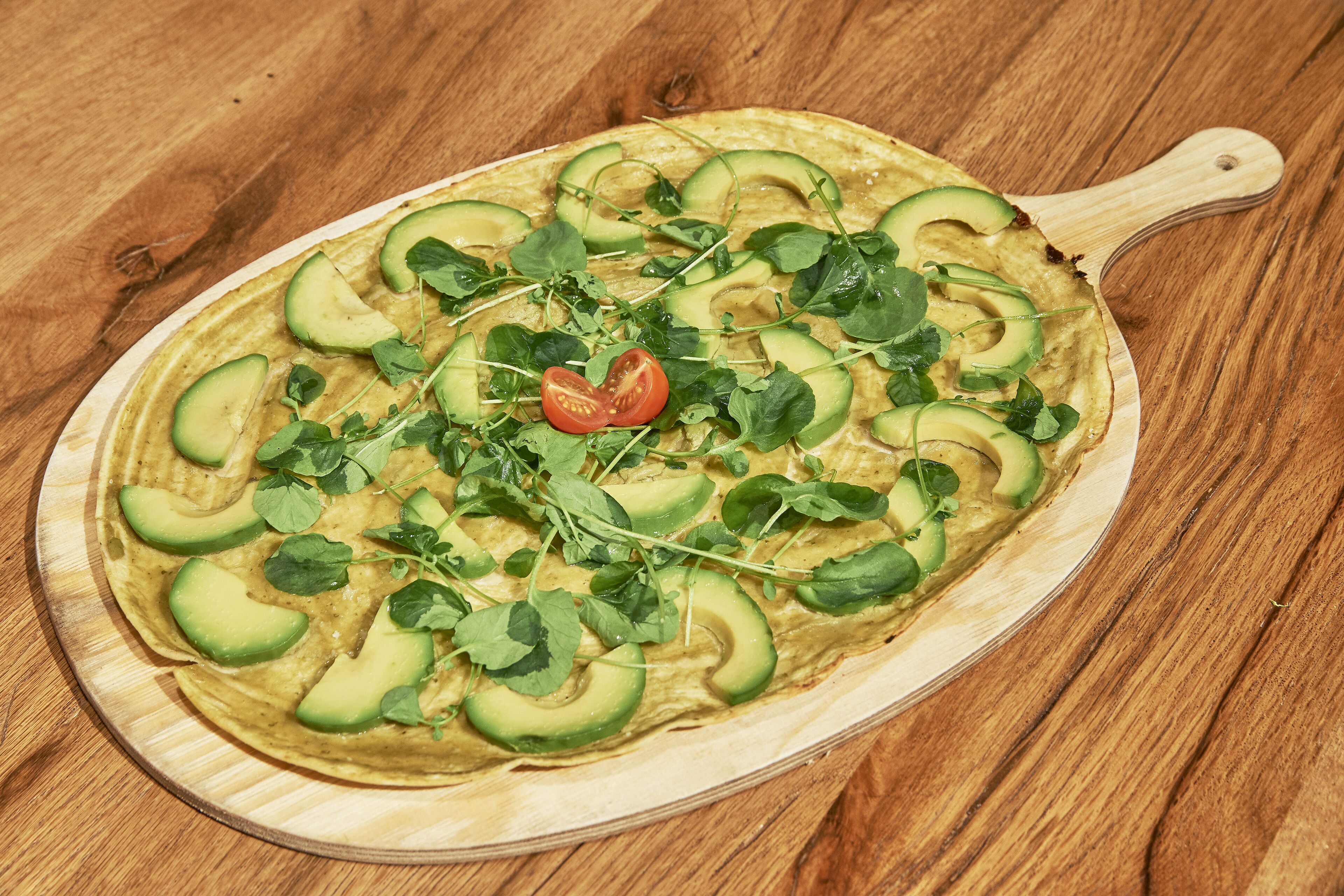 Een houten pizzabord met een pizza belegd met avocado, groen en tomaat.