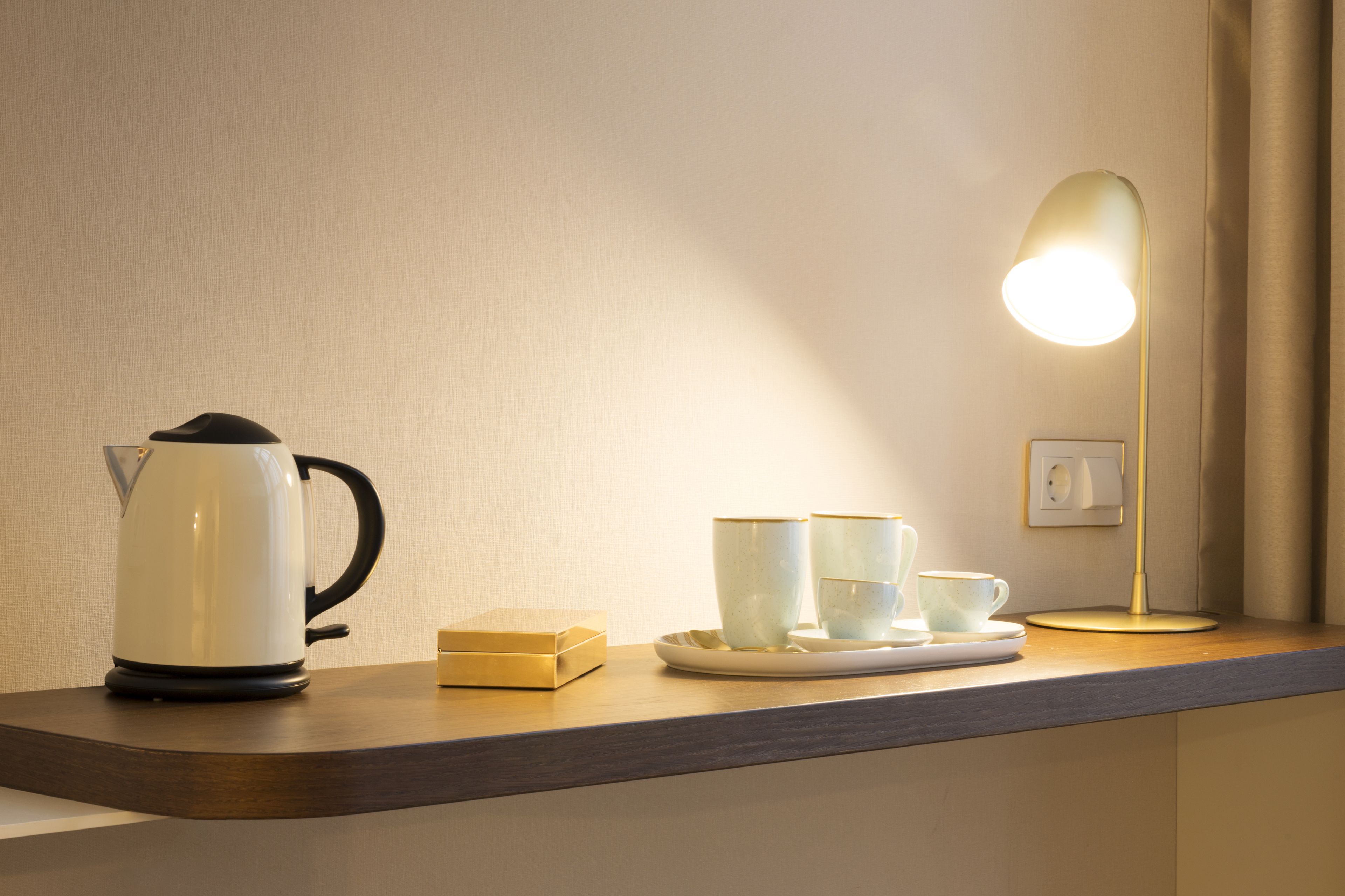A countertop with a kettle, cups, saucer, and a lamp.