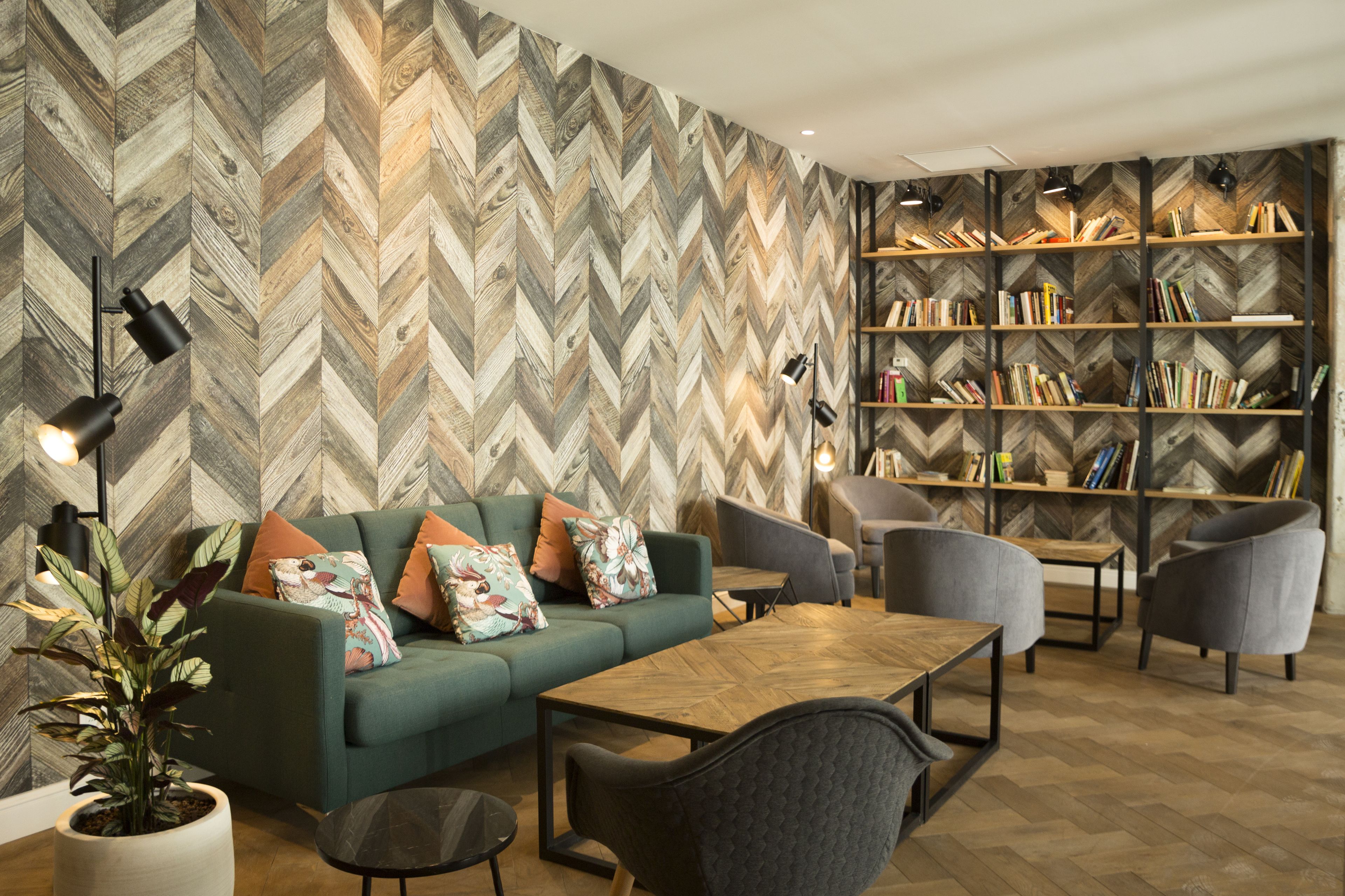 A modern living room with a herringbone-patterned wall, a green sofa with colorful cushions, a wooden coffee table, and a bookshelf filled with books.