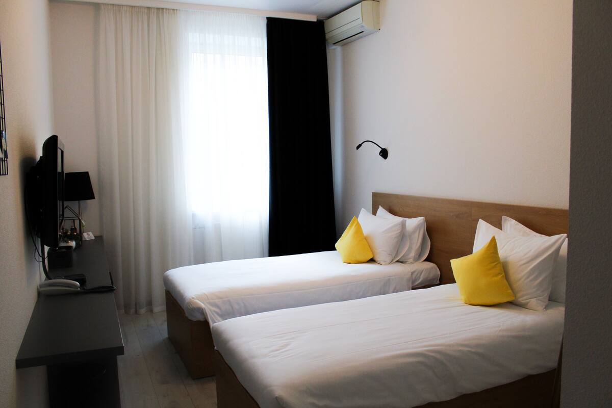 Quarto de hotel com duas camas, uma escrivaninha, uma cadeira, um abajur e uma janela com cortinas.