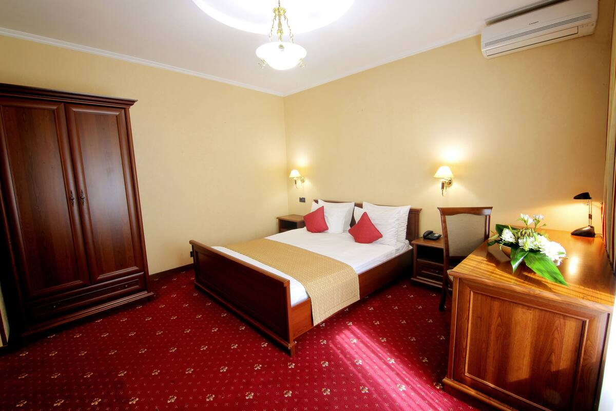 Quarto de hotel com cama grande, guarda-roupa de madeira, mesas de cabeceira e escrivaninha com abajur.