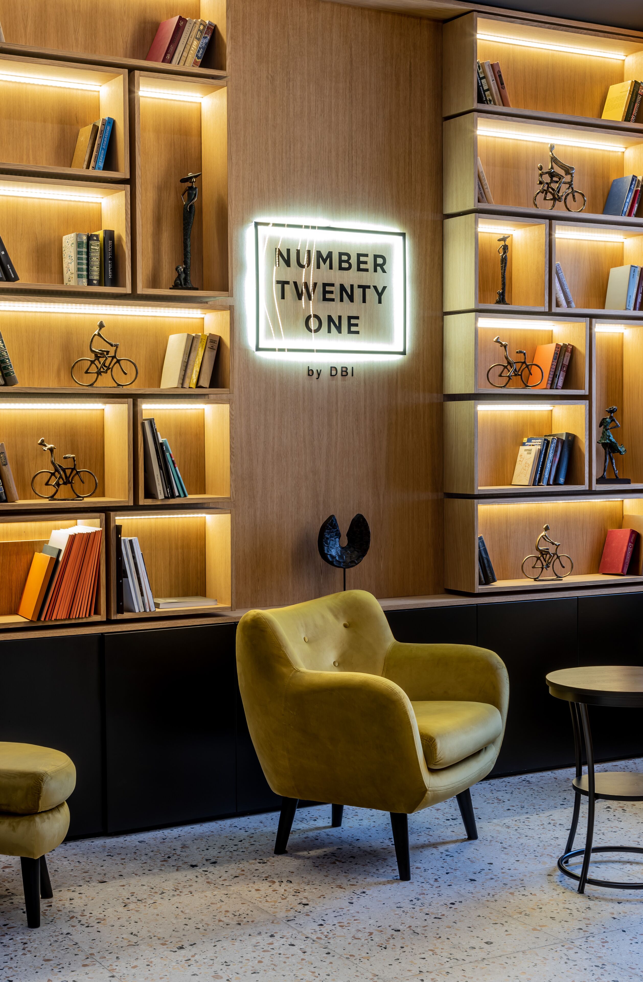 Uma biblioteca moderna com estantes de livros, uma poltrona amarela e uma placa com a inscrição 'NUMBER TWENTY ONE by DBI'.