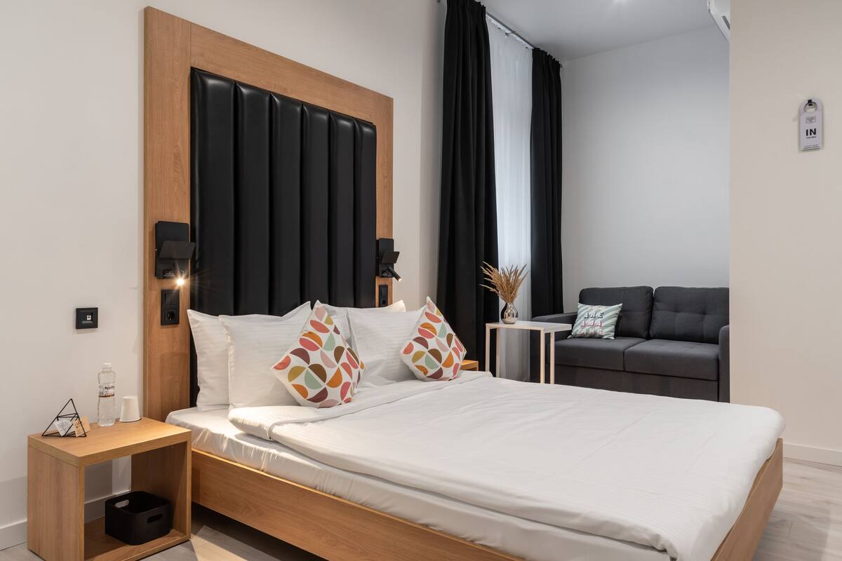 Quarto de hotel moderno com cabeceira de madeira, cama com roupa de cama branca, mesinha de cabeceira com abajur e um sofá cinzento ao fundo.