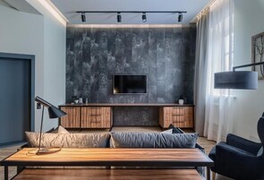 Een moderne woonkamer met een grote flatscreen televisie aan een gestructureerde muur, houten kasten eronder, een bank met kussens, een salontafel en een vloerlamp met zwart snoer.