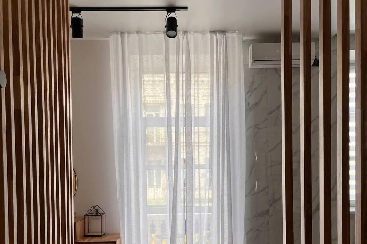 Quarto moderno com divisória de ripas de madeira, um banco, cómoda de madeira e uma janela grande com cortinas leves.