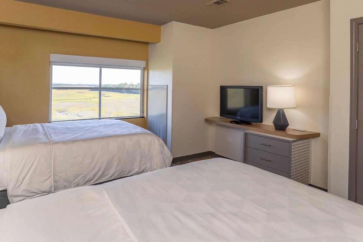 A hotel room with two beds, a television, a lamp, and a window with a view of a field.
