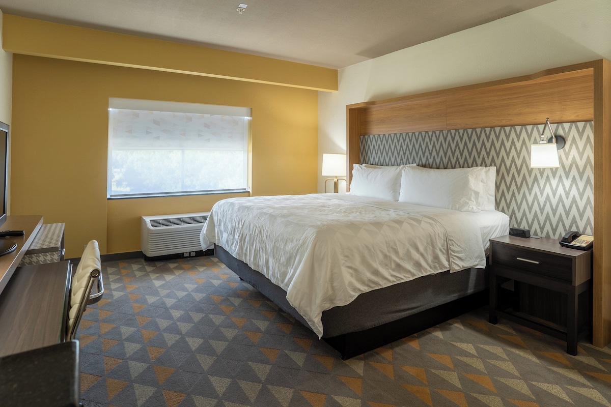 A hotel room with a bed, bedside table, lamp, and a window with blinds.