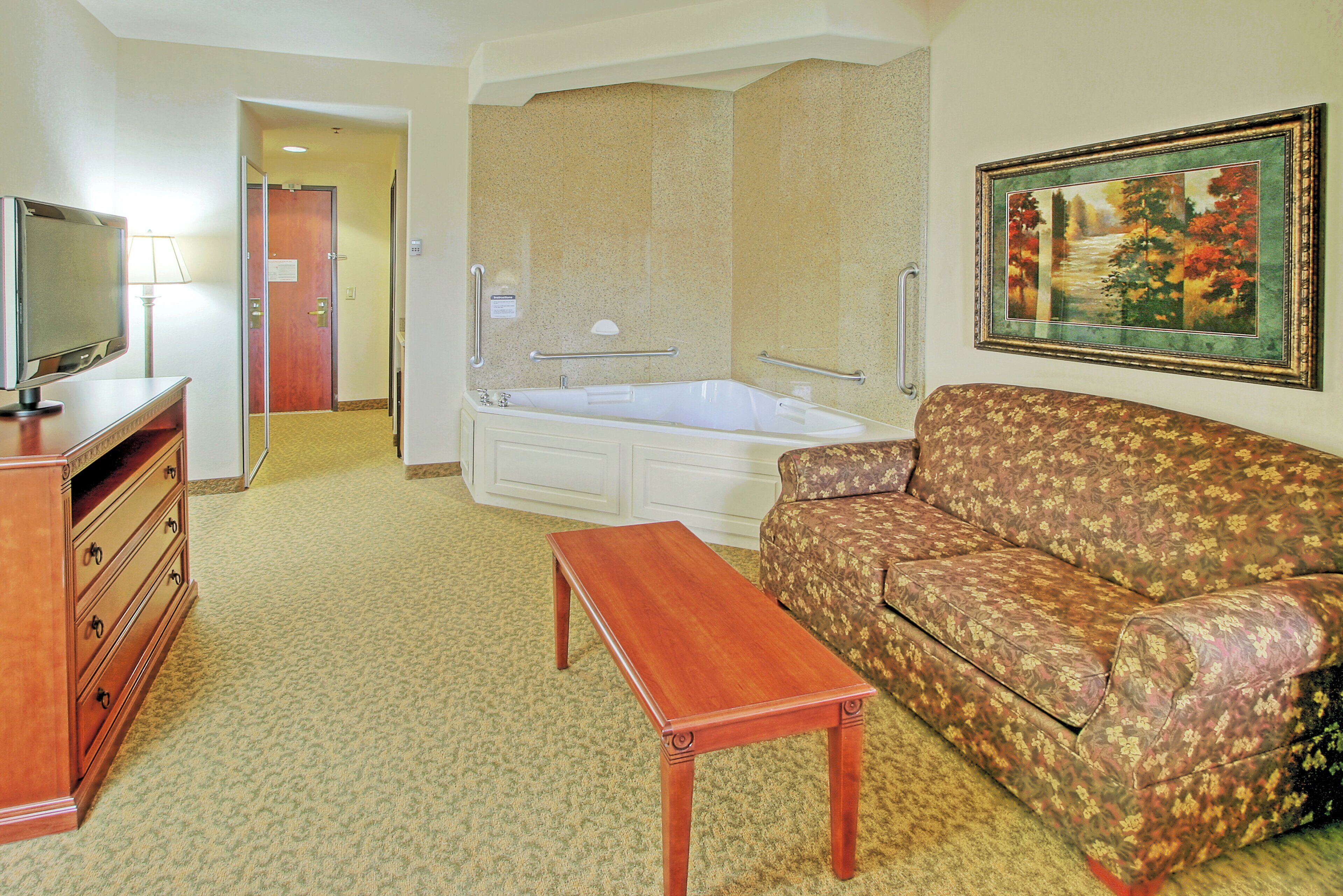 A hotel room with a TV, a sofa, a small table, a bathtub, and a painting on the wall.