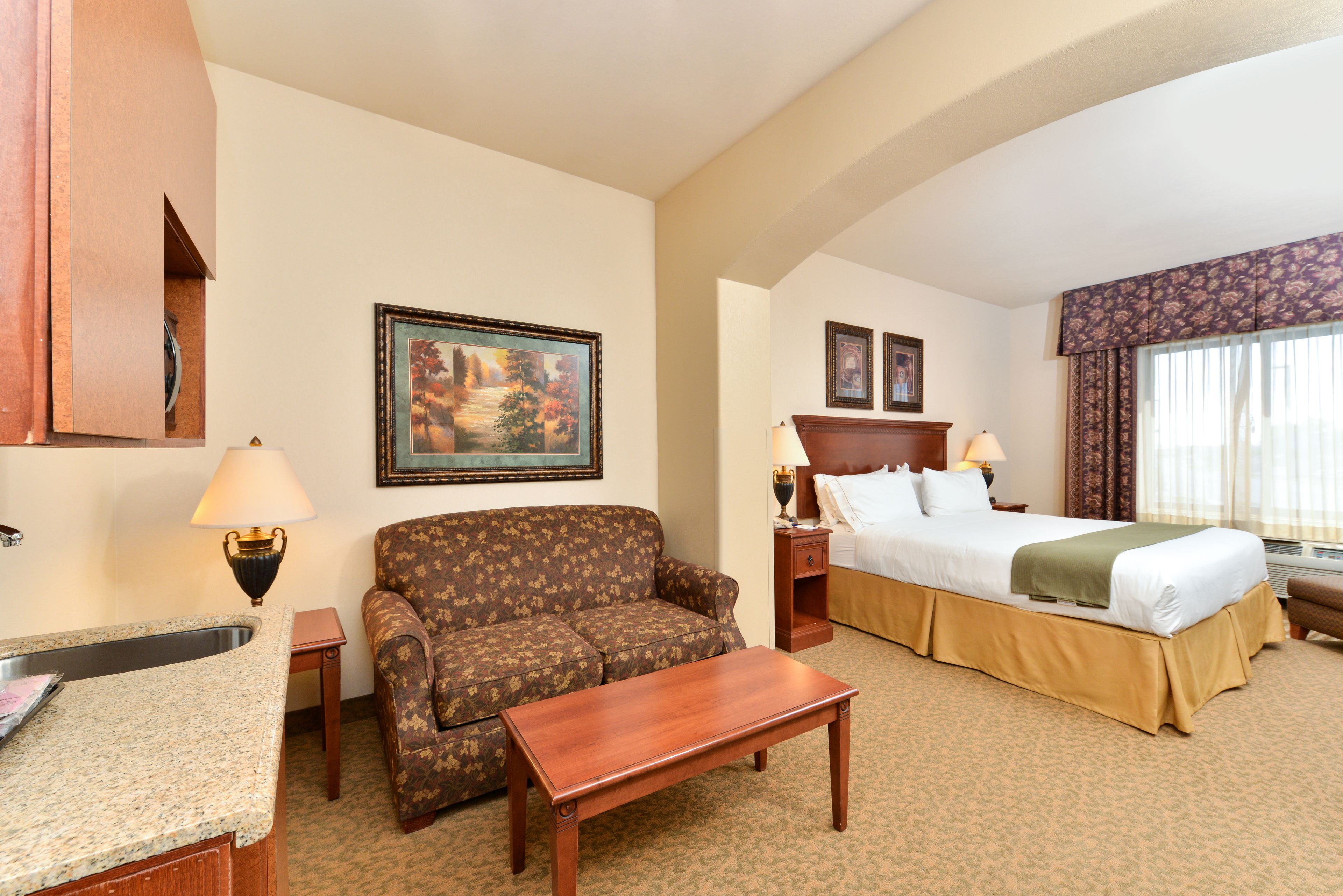 A hotel room with a bed, a sofa, a small table, a nightstand, a lamp, a painting, and a window with curtains.
