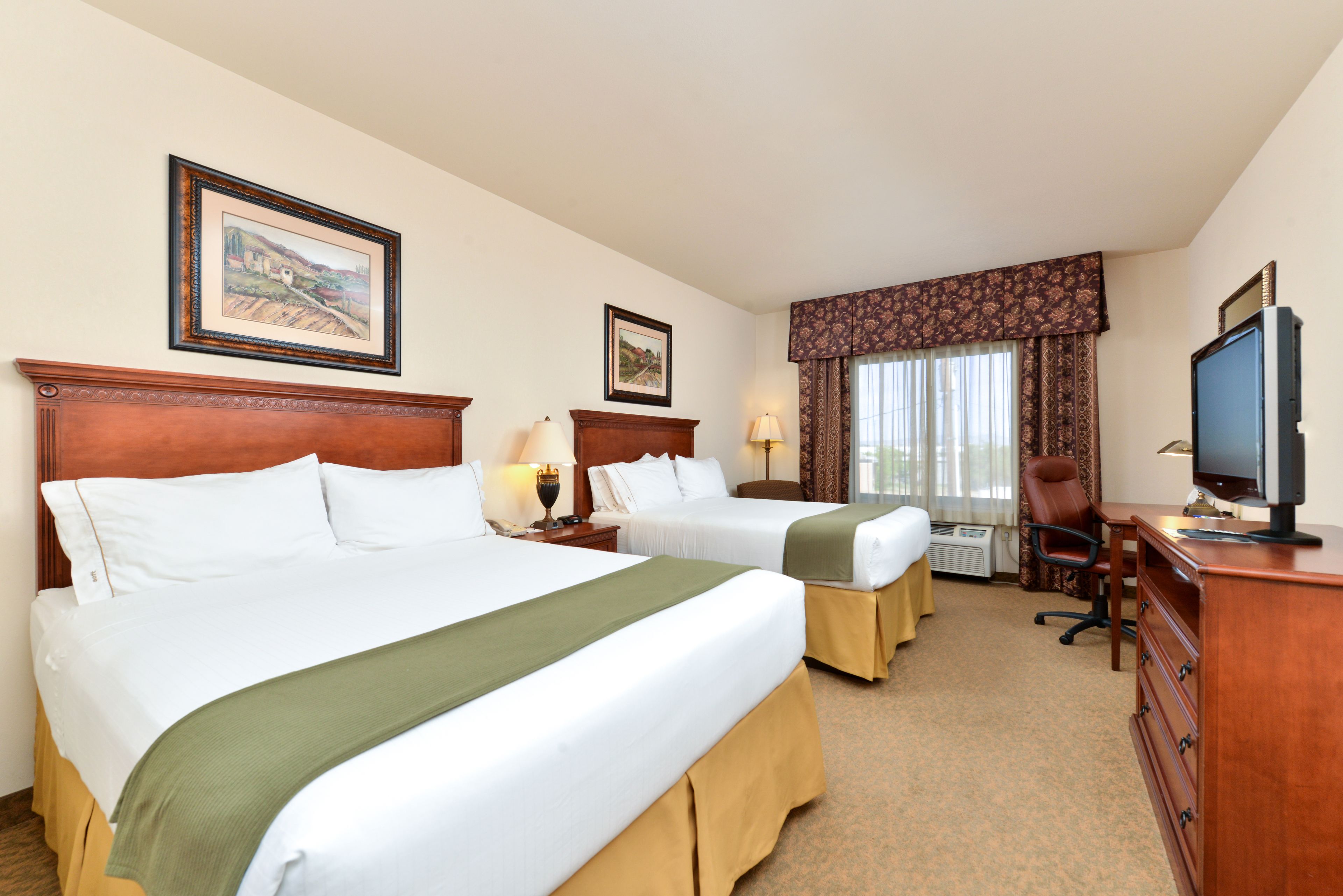 A hotel room with two beds, a desk, a chair, a television, and framed pictures on the wall.