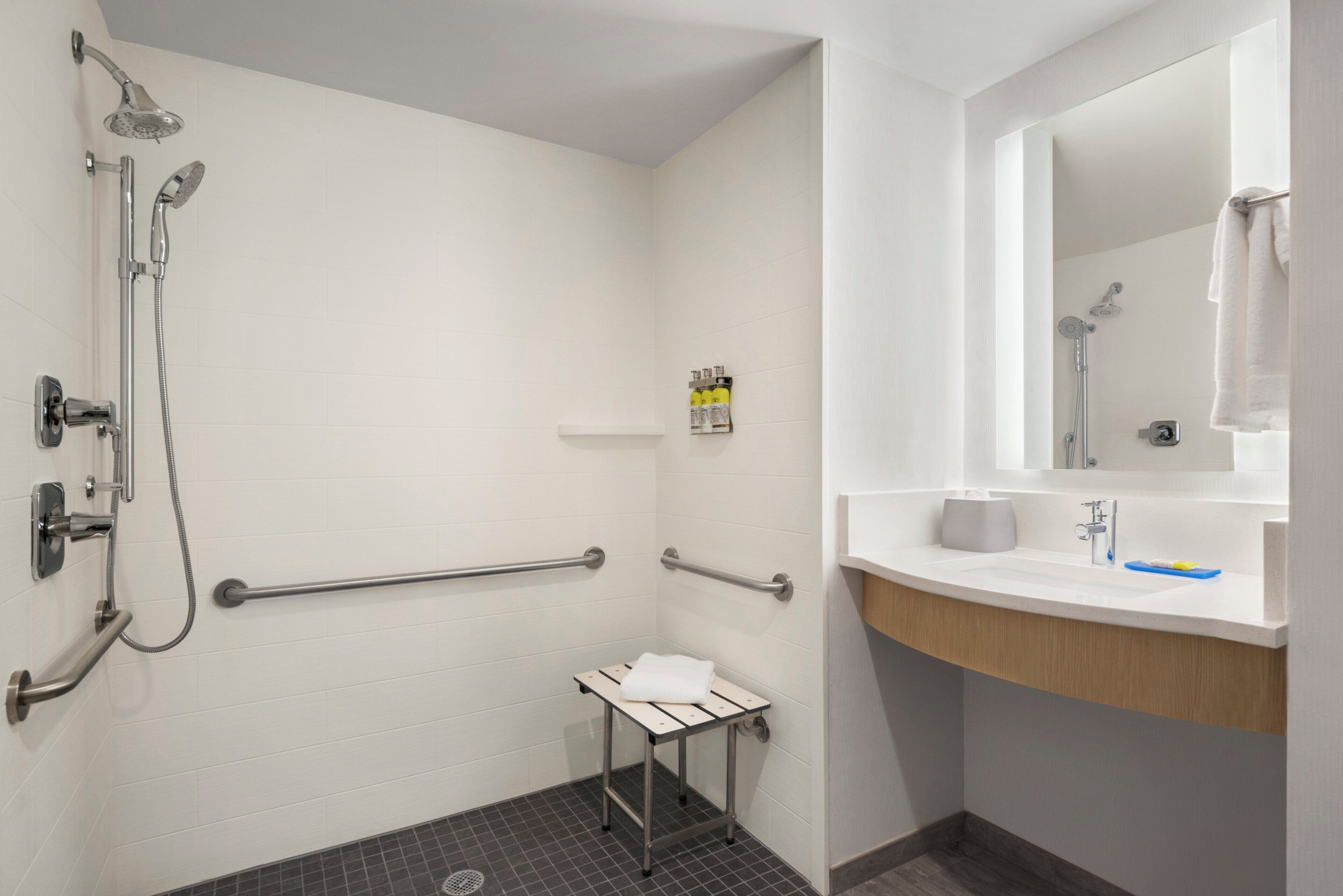 Een badkamer met een douche, handgrepen, een klein tafeltje met een handdoek, een wastafel met spiegel en toiletartikelen.