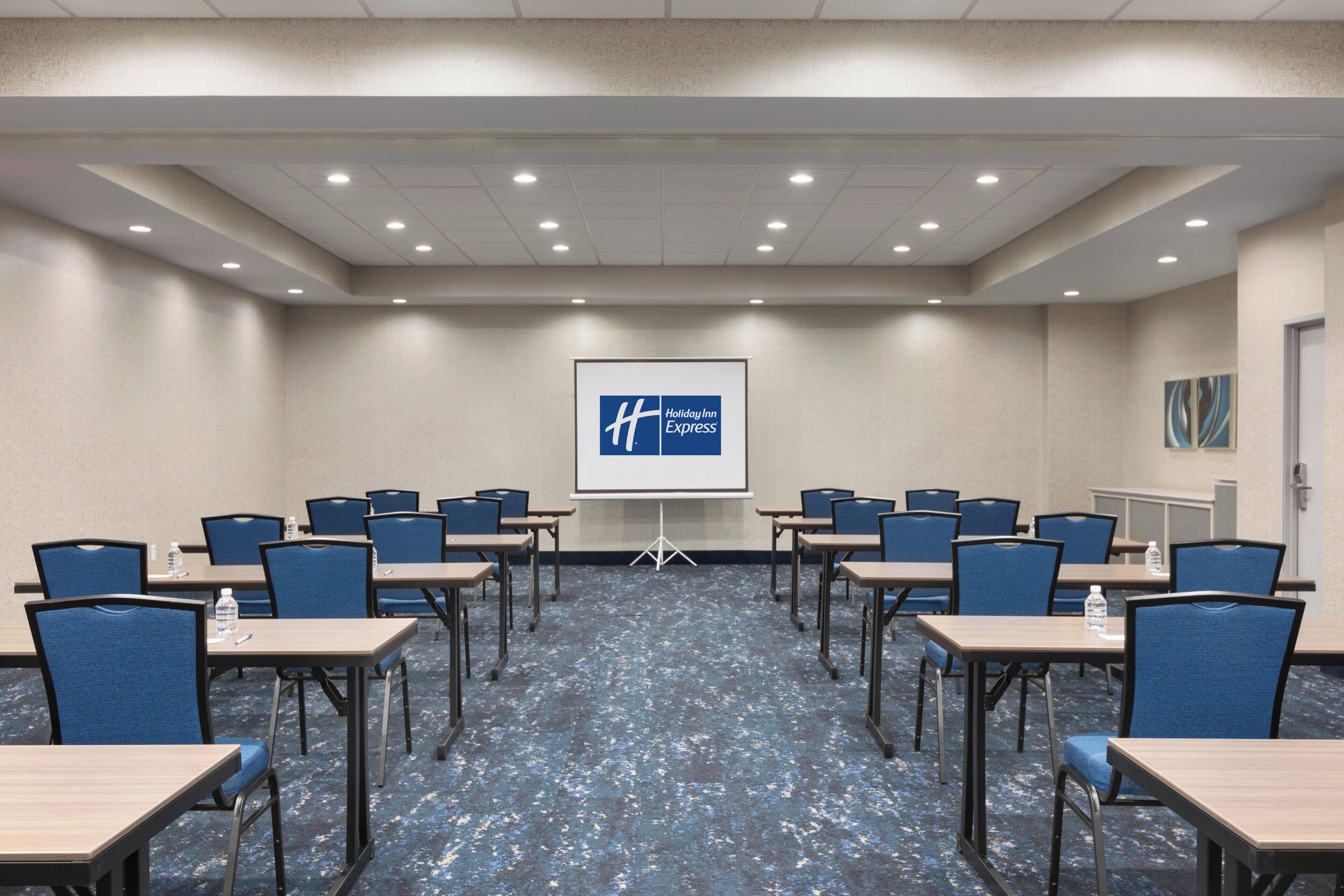A conference room with a presentation screen displaying the Holiday Inn Express logo, arranged tables with chairs, and a carpeted floor.