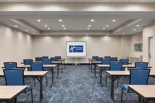 Un salón de conferencias con una pantalla de presentación que muestra el logotipo de Holiday Inn Express, mesas dispuestas con sillas y suelo alfombrado.