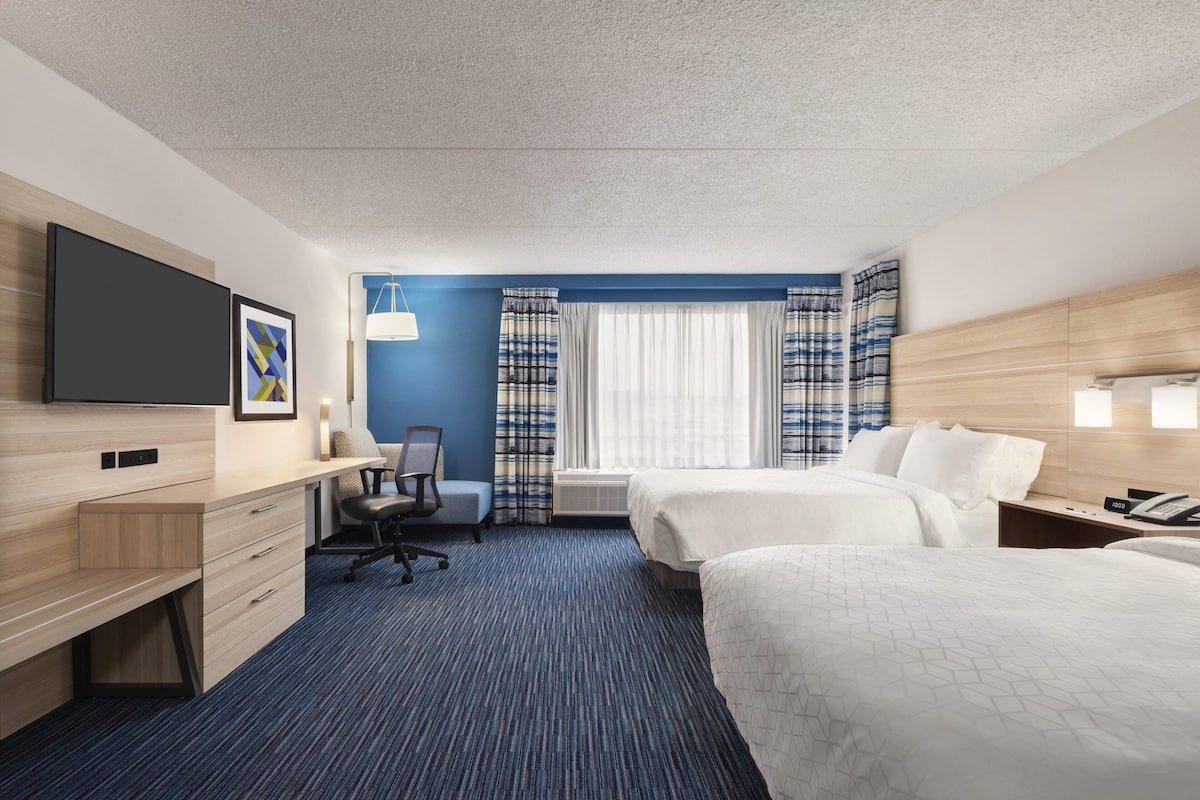 A hotel room with a desk, two beds, a chair, a flat-screen TV, and a window with curtains.