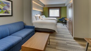 A hotel room with a blue sofa, a wooden coffee table, two beds, a desk, and a window. - Holiday Inn Express & Suites West Palm Beach Metrocentre by IHG (West Palm Beach)