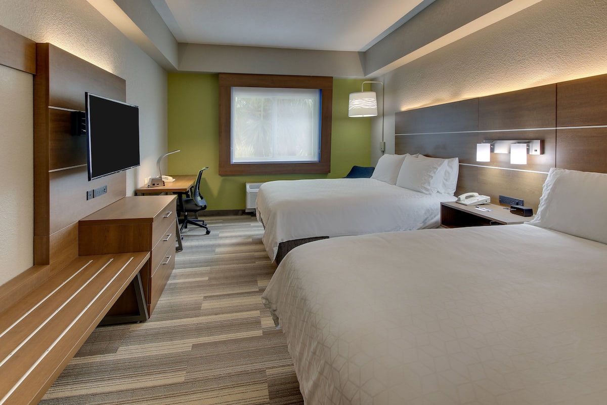 Una habitación de hotel con dos camas, un escritorio con silla, un televisor de pantalla plana, una ventana y una lámpara fijada a la pared.