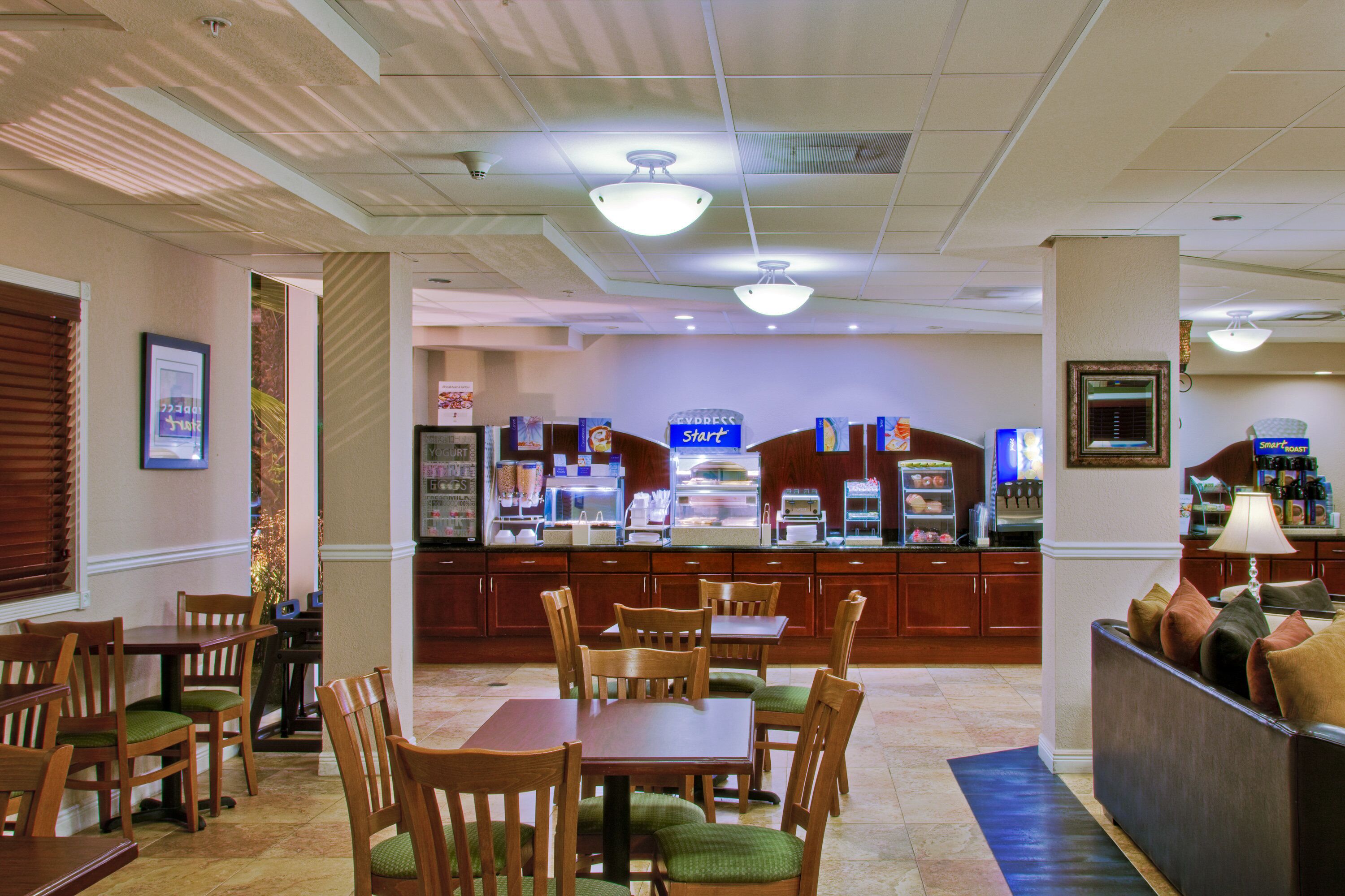 A hotel dining area with wooden tables and chairs, a buffet counter, and a seating area with cushions.