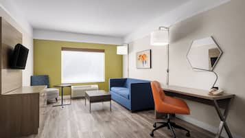 A modern hotel room with a wooden desk, a blue sofa, an orange chair, a small table, and a flat-screen TV.