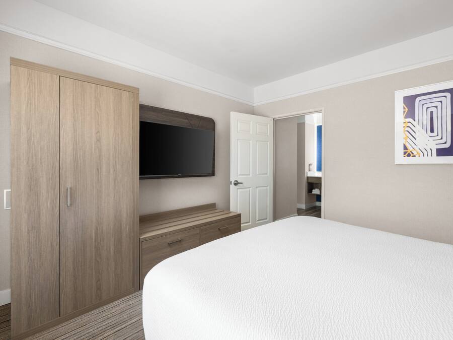 Habitación de hotel con una cama grande, un armario, un televisor fijado a la pared y una puerta que da a otra habitación.