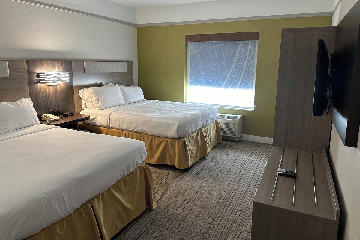 A hotel room with two beds, a flat-screen TV, a bedside table with a lamp, a window with a view, and a wall-mounted heater.