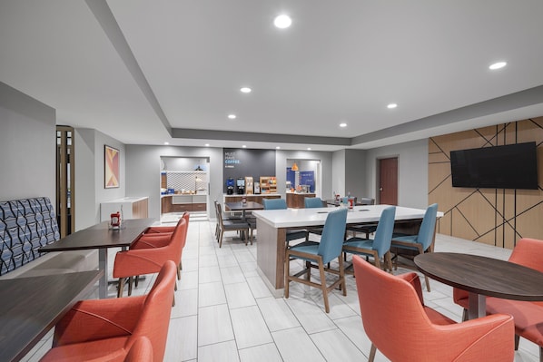 A modern dining area with a long table and orange chairs, a smaller table with blue chairs, and a TV mounted on the wall. - Holiday Inn Express Hotel & Suites San Dimas by IHG (San Dimas)