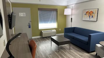 A modern hotel room with a wooden desk, a blue sofa, a small table, and a large window with a view.