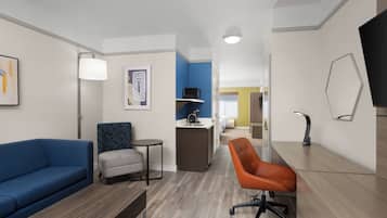 A hotel room with a blue sofa, a grey armchair, an orange office chair, a small round table, a desk, a lamp, a flat-screen TV, and a bed in the background.
