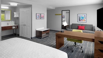 A hotel room with a bed, a desk with a chair, a grey sofa with a red pillow, a mirror, and artwork on the walls.