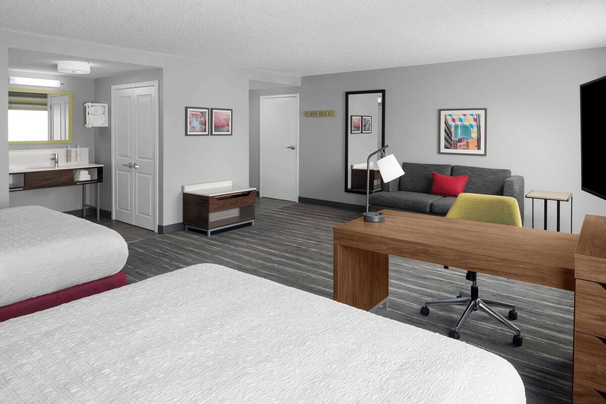 Ein Hotelzimmer mit zwei Betten, einem Schreibtisch, einem grauen Sofa mit roten und gelben Kissen, einem Spiegel und gerahmten Bildern an den Wänden.