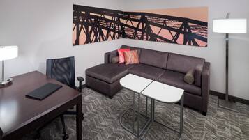 Una habitación de hotel moderna con un sofá modular, una mesita pequeña, un escritorio con una silla, una lámpara y un cuadro abstracto grande en la pared.