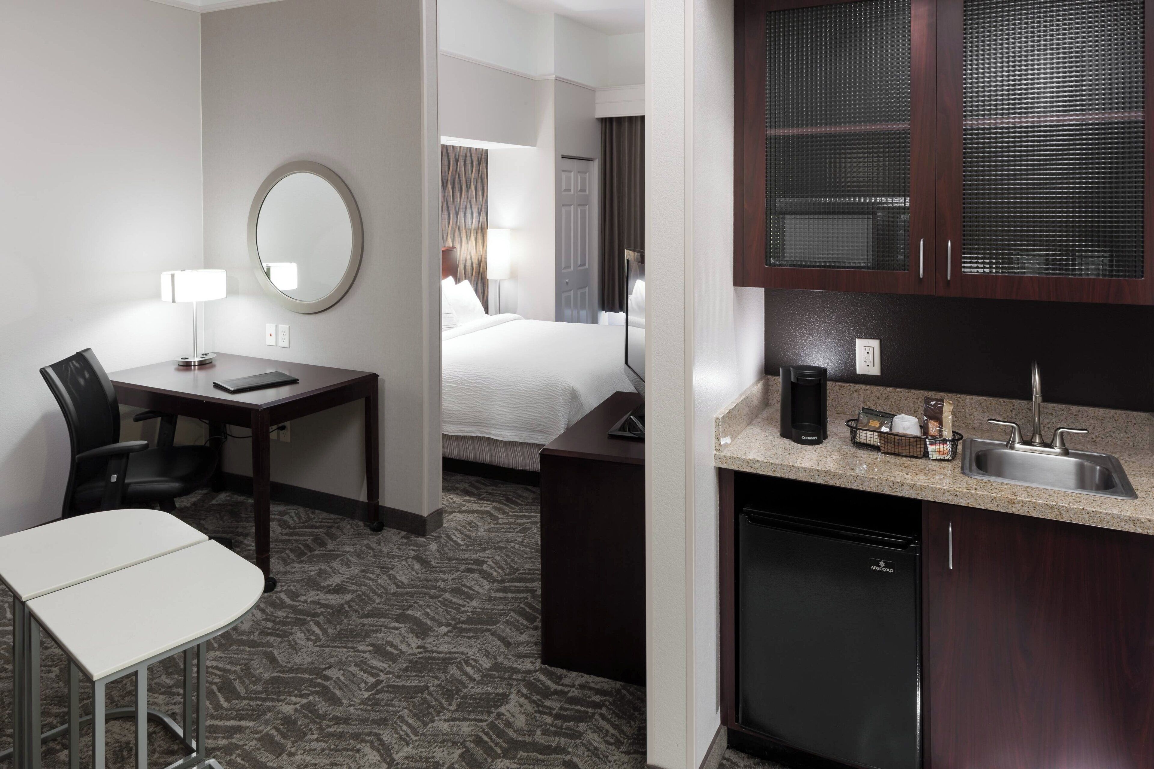 Ein Hotelzimmer mit einem Bett, einem Schreibtisch mit Stuhl, einer kleinen Küchenzeile, einem runden Spiegel und einem durchgegangene Tür sichtbaren Badezimmer.