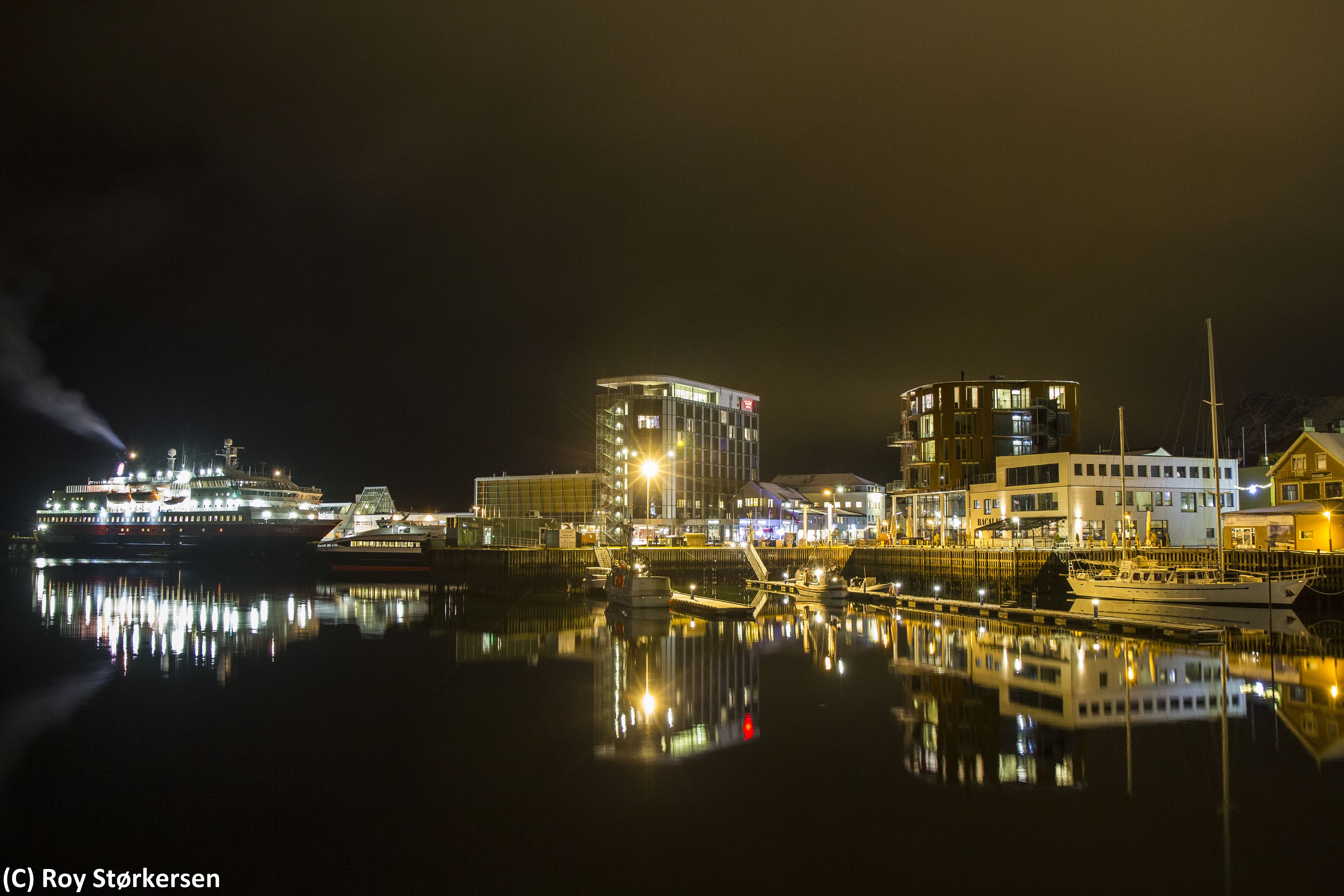 Paisaje urbano frente al agua con edificios iluminados y un crucero amarrado en el puerto.
