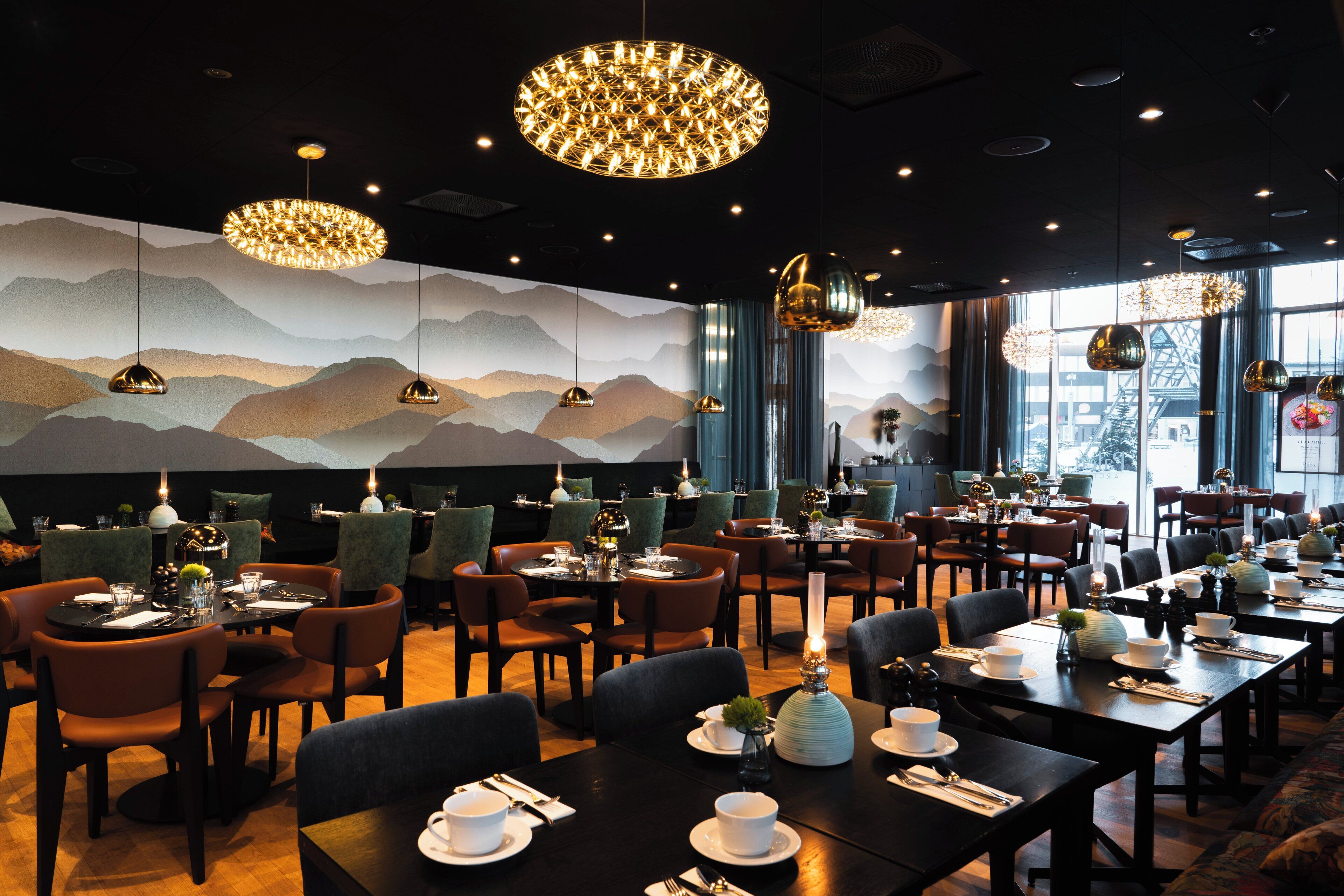 A restaurant with multiple tables and chairs, a mountain-themed wall, and hanging pendant lights.