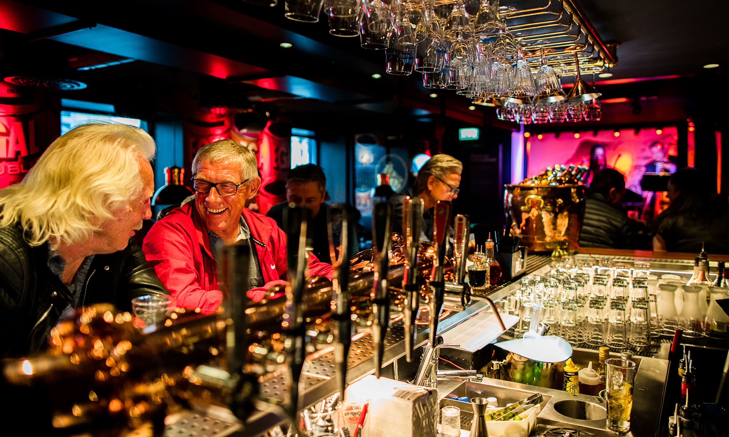 Zona de bar con varios vasos y botellas, una barra y una persona con chaqueta roja.