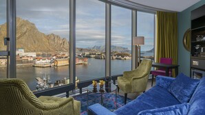 Habitación moderna con un amplio ventanal que ofrece vistas a un fiordo, barcos y montañas. Hay un sofá azul, un sillón verde estampado y una pequeña mesa redonda con objetos decorativos.