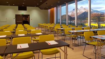 Een congreszaal met gele stoelen en tafels, grote ramen met uitzicht op bergen en een deur met brandalarm.