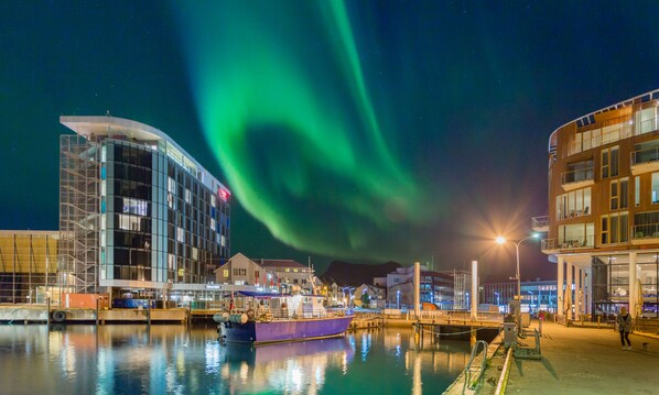 Un edificio moderno frente al agua, un muelle con un bote y la aurora boreal en el cielo.