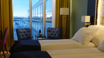 A hotel room with a large bed, two armchairs, a small table, and a view of a cityscape through large windows.