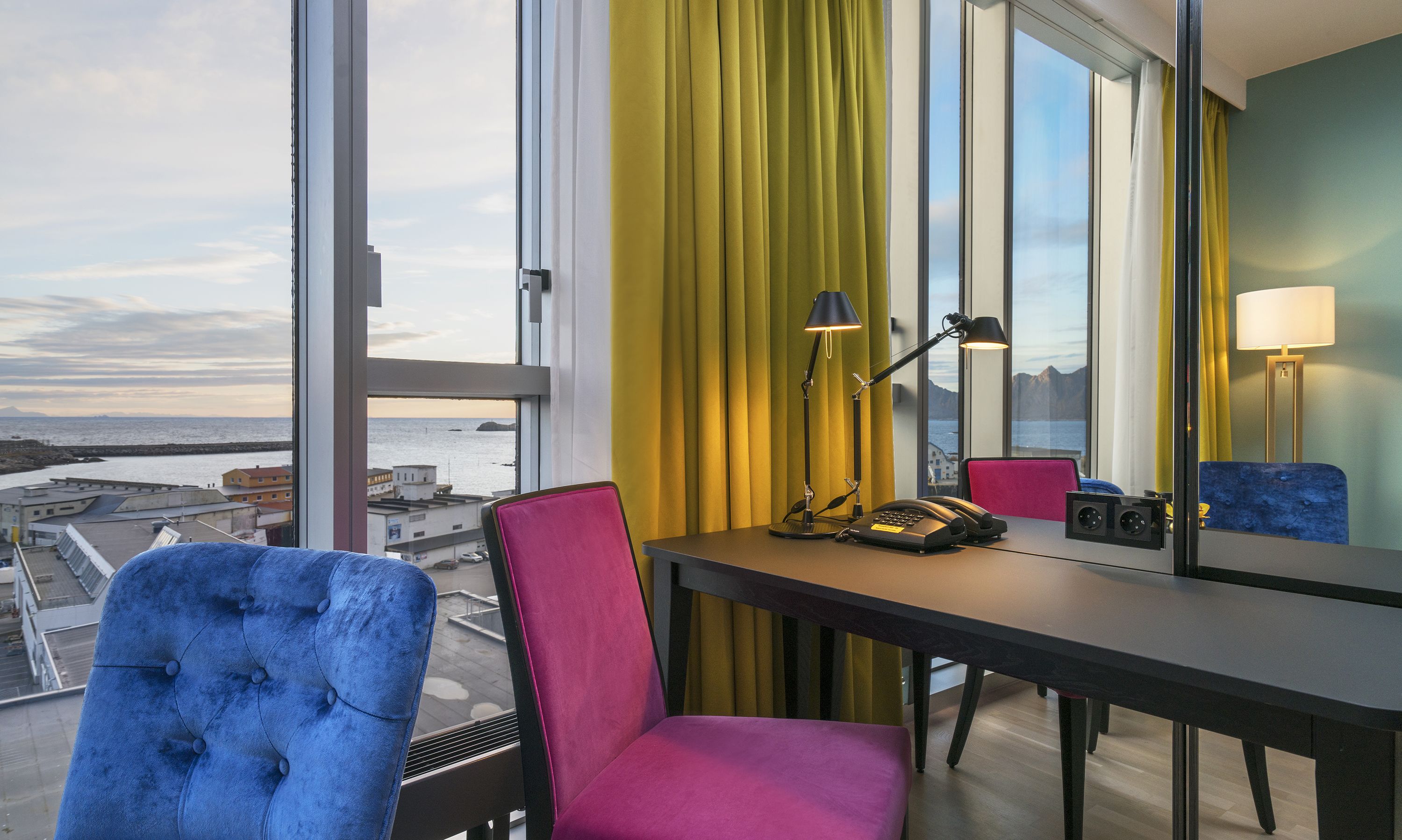 A hotel room with a view of the sea, a desk with a telephone and lamp, and two chairs.