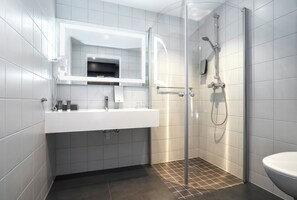 Un baño moderno con ducha acristalada, un lavamanos blanco con espejo y suelo oscuro.