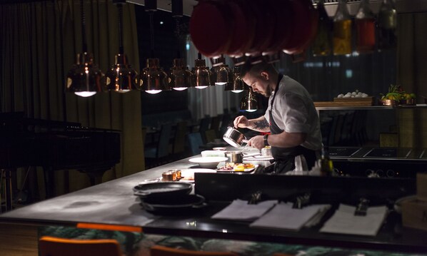 Un chef prepara comida en una cocina tenuemente iluminada con lámparas colgantes sobre la encimera.