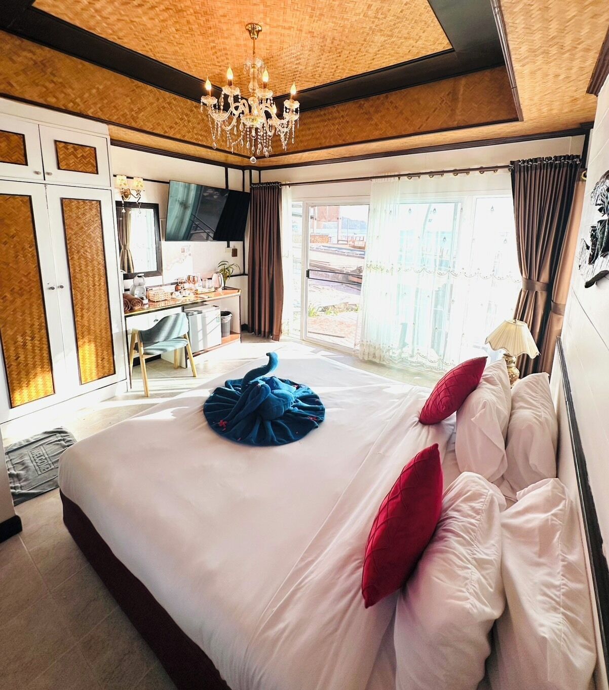 A hotel room with a large bed, a desk, a chair, a chandelier, and a view of the outdoors.