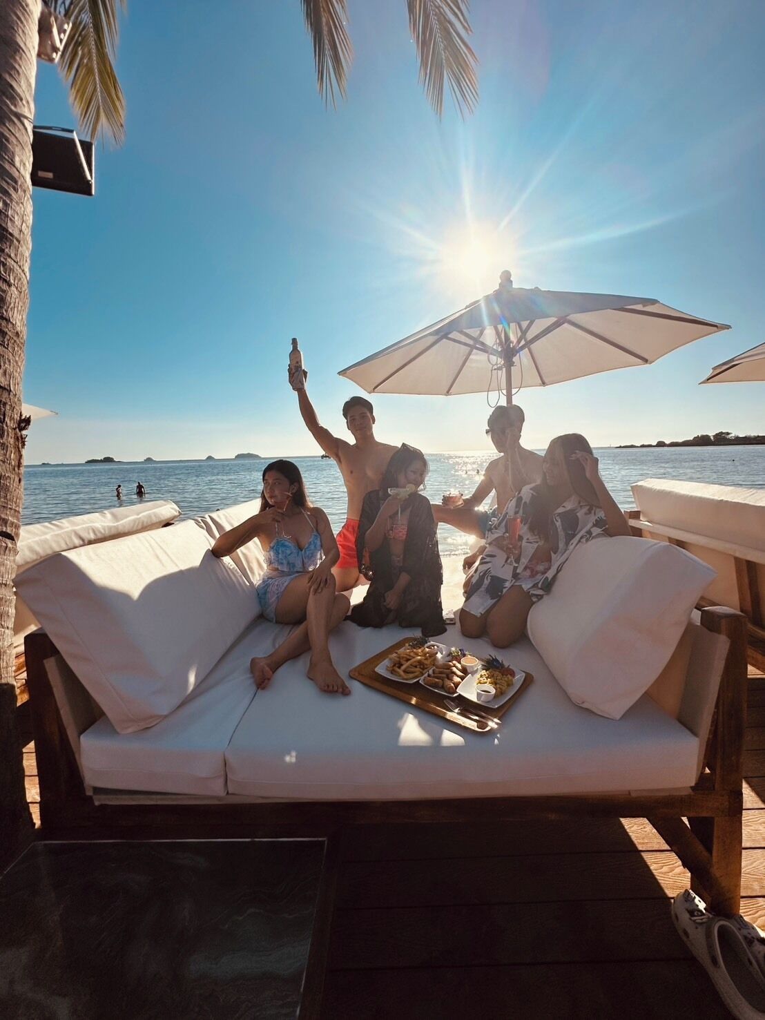 A group of people relaxing on a white outdoor sofa with a wooden table and a tray of food, overlooking a beach with palm trees and the ocean.